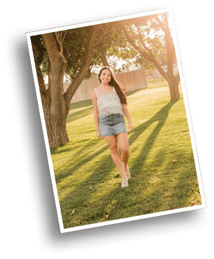 A person with long hair wearing a sleeveless top and denim skirt is walking on grass in a sunlit park with large trees casting long shadows. Description generated by AI