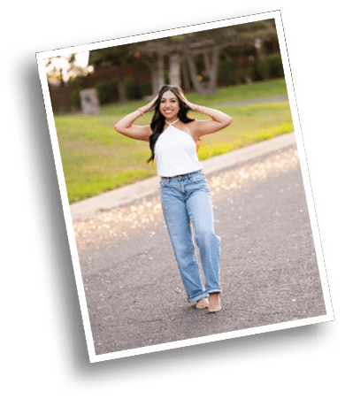 A person with long hair wearing a white sleeveless top and blue jeans is standing on a paved path with hands touching their head, smiling, with trees and grass in the background. Description generated by AI