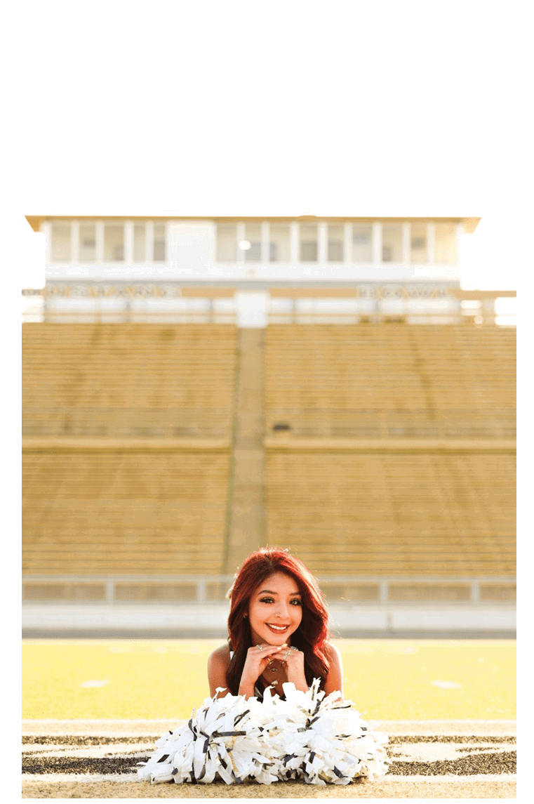 A person with long red hair is smiling and resting their chin on their hands behind white pom-poms on a sports field with empty bleachers in the background. Description generated by AI