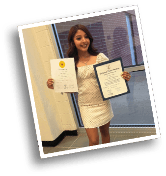 A person wearing a light-colored dress with black polka dots is holding certificates while standing indoors near a window and a tiled floor. Description generated by AI