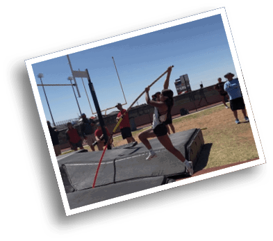 A person is mid-air attempting a pole vault jump over a padded landing area while two people in red shirts observe in the background. Description generated by AI