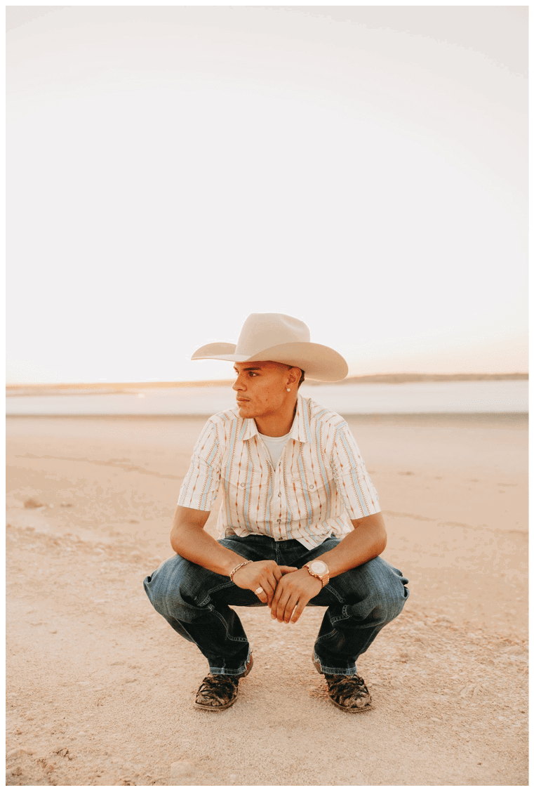 A person wearing a wide-brimmed hat, striped short-sleeve shirt, jeans, and boots is squatting on a sandy beach near calm water during sunset. Description generated by AI