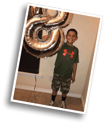 A smiling child wearing a green shirt with an orange logo and camouflage shorts is holding a large silver balloon shaped like the number three. Description generated by AI