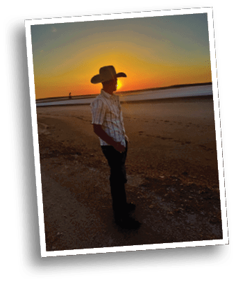 A person wearing a cowboy hat and a striped shirt stands on a beach at sunset with footprints visible in the sand. Description generated by AI