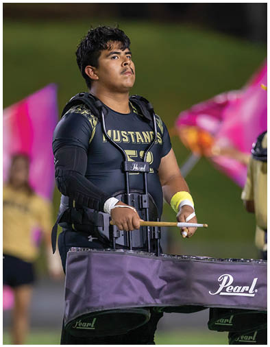 A person wearing a Mustangs shirt and protective gear is playing a percussion instrument with drumsticks outdoors, with blurred people and colorful flags in the background. Description generated by AI