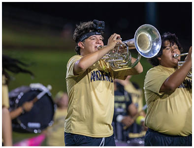 People wearing yellow shirts are playing brass instruments and drums in an outdoor band performance at night. Description generated by AI