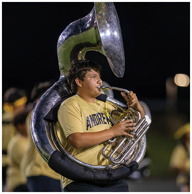A person wearing a yellow shirt with the word “ANDREWS" is playing a large silver sousaphone outdoors at night. Description generated by AI