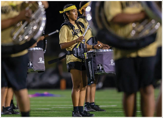 A person wearing a yellow shirt and headband is playing a snare drum in a marching band on a field at night. Description generated by AI