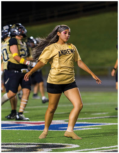 A person with long hair tied back is wearing a yellow sports jersey with the word “ANDREWS" and appears to be in motion outdoors at night. Description generated by AI