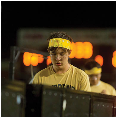 A person with braided hair and glasses is looking down with a focused expression against a blurred background of people sitting on bleachers. Description generated by AI