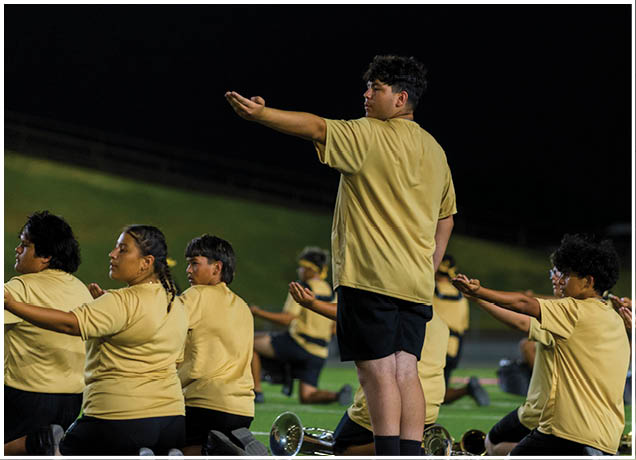 A group of people wearing matching yellow shirts and black shorts are kneeling on a field at night, extending one arm forward in a coordinated pose. Description generated by AI