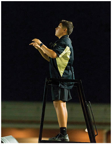 A person wearing a black and yellow shirt is standing against a dark background with their hands raised as if conducting or directing. Description generated by AI