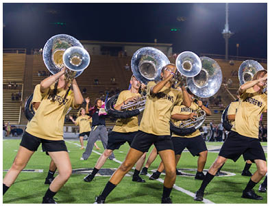 People wearing matching yellow shirts and black shorts are playing sousaphones while performing a coordinated routine on a football field at night. Description generated by AI
