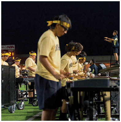 A group of people wearing matching yellow shirts and headbands are playing percussion instruments outdoors at night while a person stands on a platform conducting them. Description generated by AI