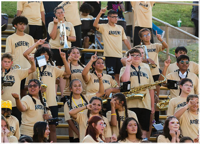 A group of people wearing matching yellow shirts with “ANDREWS" printed on them are sitting on bleachers playing musical instruments and raising their index fingers. Description generated by AI
