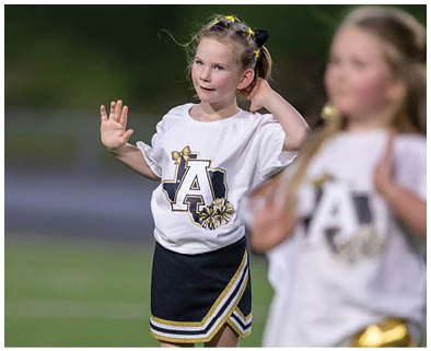 A child in a cheerleading outfit with a white shirt and dark skirt is standing on a field with one hand raised and the other touching their head while another child in similar attire is blurred in the foreground. Description generated by AI