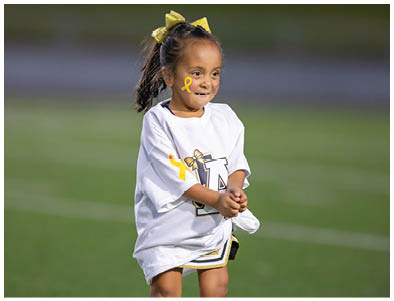 A child wearing a white sports jersey and shorts is standing on a grassy field with hands clasped together. Description generated by AI