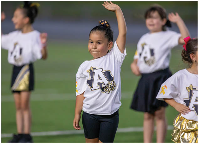 A child wearing a white shirt with a large letter A and decorative bows is raising one arm while standing on a field with other children in similar outfits. Description generated by AI