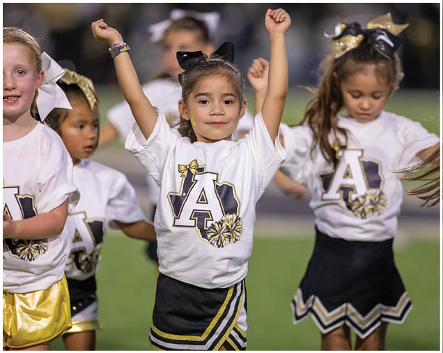 Children wearing cheerleading outfits with large bows and holding their arms up are performing on a field. Description generated by AI