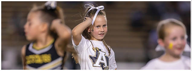 Children wearing cheerleading and sports uniforms are standing on a field, with one child raising an arm. Description generated by AI