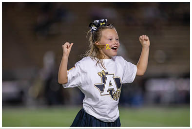A child wearing a cheerleading outfit with a large bow in their hair and a yellow ribbon on their cheek is cheering with raised fists on a sports field. Description generated by AI