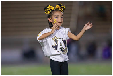 A child with braided hair and yellow and black bows is wearing a white shirt with a large letter A and holding a small yellow ribbon near their face while standing outdoors. Description generated by AI