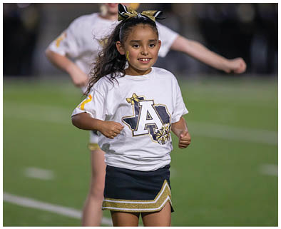 A child wearing a cheerleading outfit with a white shirt and dark skirt is smiling and performing on a sports field. Description generated by AI