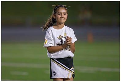 A child wearing a cheerleading outfit stands on a sports field with hands clasped and a focused expression. Description generated by AI