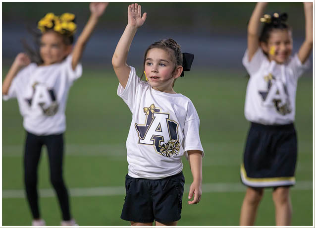 A child wearing a white cheerleading shirt with a large letter A and a bow design is raising one hand while standing on a field with other children in similar outfits. Description generated by AI