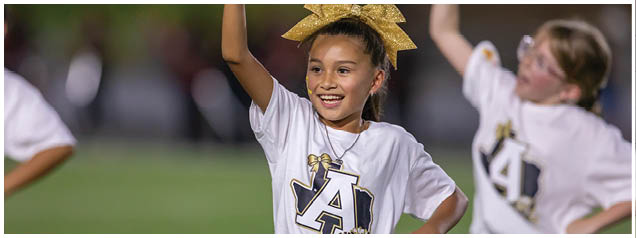 A child wearing a white shirt with a large letter A and a gold bow in their hair is smiling and raising one arm while another child in a similar outfit is blurred in the background. Description generated by AI