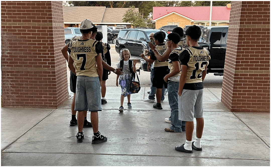 A group of people wearing sports jerseys and casual clothing are standing and talking under a covered brick walkway near parked cars and trees. Description generated by AI