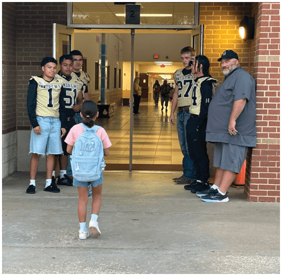 A group of people wearing sports jerseys are standing and talking near a school hallway entrance while a child with a backpack stands facing them. Description generated by AI