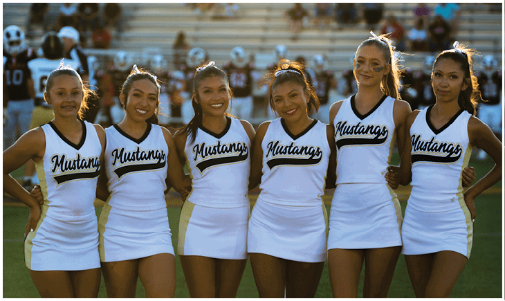 Six adults wearing matching white cheerleading uniforms with “Mustangs" written on the front stand closely together on a sports field with a blurred crowd and players in the background. Description generated by AI