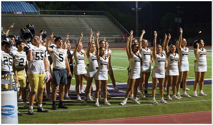 A group of people in sports uniforms and cheerleading outfits stand on a football field at night with their arms raised. Description generated by AI