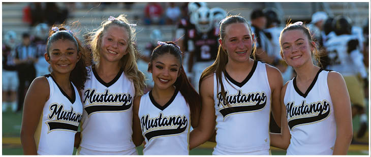 Five people wearing white cheerleading uniforms with “Mustangs" written on them are standing closely together and smiling on a sports field. Description generated by AI