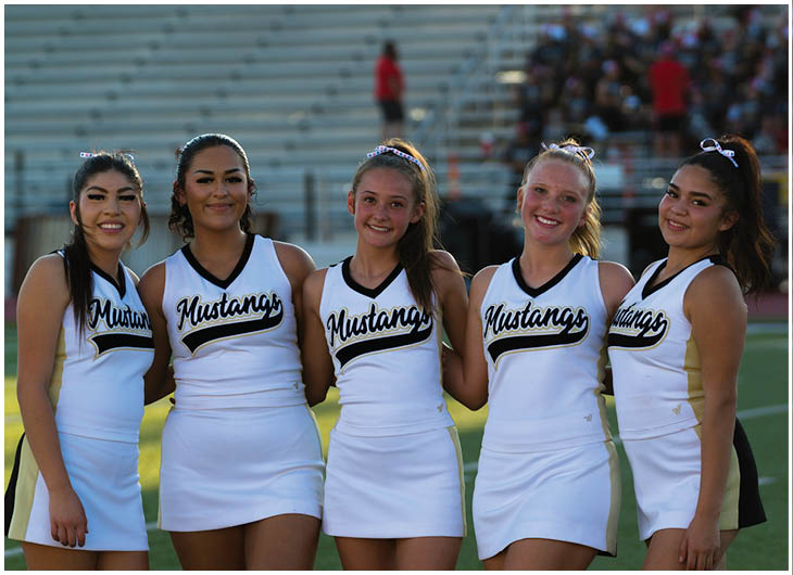 Five people wearing matching white cheerleading uniforms with “Mustangs" written on the front stand closely together on a sports field with bleachers and a crowd in the background. Description generated by AI
