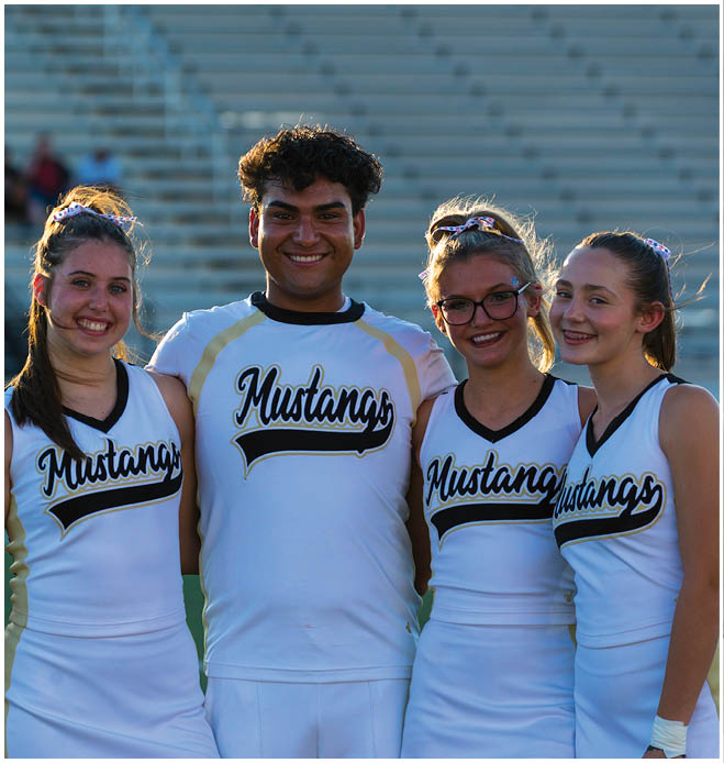 Three smiling adults wearing white cheerleading uniforms with “Mustangs" written on them stand closely together on a sports field with empty bleachers in the background. Description generated by AI