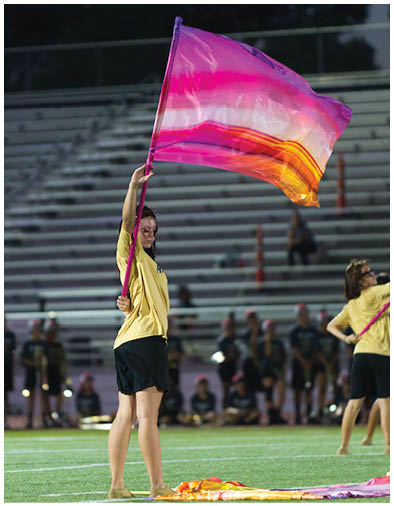 A person wearing a yellow shirt and black shorts is standing on a field holding a colorful flag raised above their head with another flag lying on the ground nearby. Description generated by AI