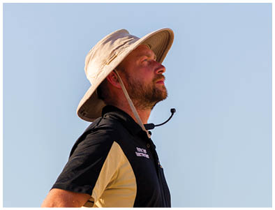 A person wearing a wide-brimmed hat and a black and beige polo shirt stands outdoors against a clear blue sky. Description generated by AI