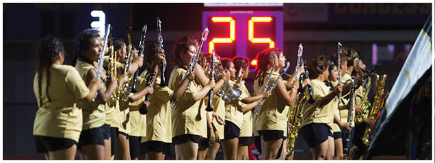 A group of people wearing matching light-colored outfits are standing in a line playing wind instruments at an outdoor event with a digital scoreboard in the background. Description generated by AI