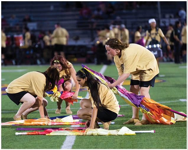 Four people wearing matching yellow shirts and black shorts are kneeling and crouching on a sports field while handling colorful flags or fabric. Description generated by AI