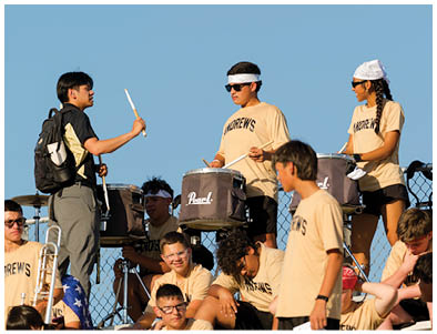 A group of people wearing matching light-colored shirts with “ANDREWS" printed on them are playing drums and other instruments outdoors against a clear blue sky. Description generated by AI