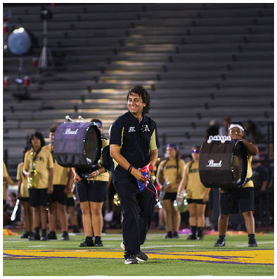 A person in a black and yellow uniform stands smiling on a football field holding drumsticks while a marching band in yellow shirts and black shorts plays instruments in the background. Description generated by AI
