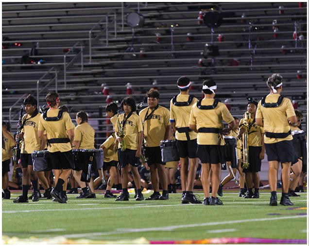 People wearing matching yellow shirts and black shorts are standing and walking on a sports field with musical instruments, with empty bleachers in the background. Description generated by AI