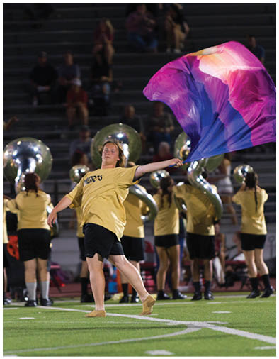 A person in a yellow shirt and black shorts is performing a flag routine on a football field with a colorful purple and pink flag while a marching band plays in the background. Description generated by AI