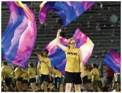 A person wearing a yellow sleeveless shirt with “ANDREWS" printed on it and black shorts is raising one arm while standing on a field with other people in similar attire holding colorful flags in the background. Description generated by AI
