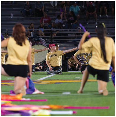 People wearing yellow shirts with “ANDREWS" printed on them are performing with musical instruments in front of seated spectators in bleachers. Description generated by AI