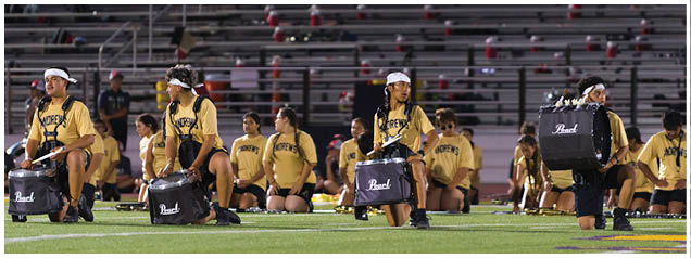A group of people wearing matching yellow shirts and white headbands are kneeling on a sports field, some holding drums while others sit behind them. Description generated by AI