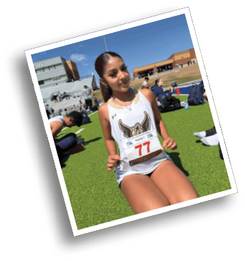 Person wearing a white athletic jersey with a winged logo and holding a race bib numbered 77 at an outdoor track and field event. Description generated by AI