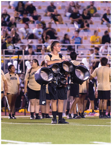 A person wearing a band uniform stands on a field holding a large percussion instrument with multiple drums while other people in similar uniforms stand and walk nearby in a stadium setting. Description generated by AI